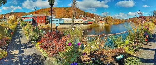 A vibrant riverside scene with colorful buildings, lush flowers, and autumn foliage under a blue sky with fluffy clouds.