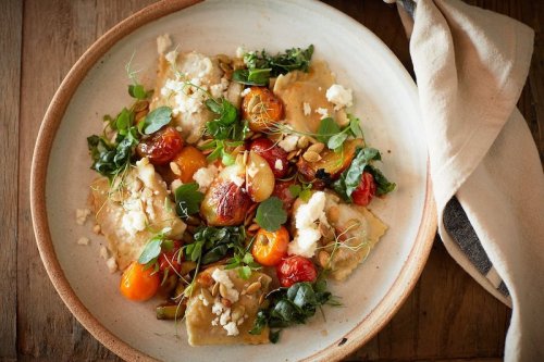 A plate of ravioli topped with cherry tomatoes, greens, and crumbled cheese, garnished with microgreens.