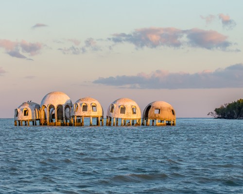 Five dome-shaped structures on stilts in calm water, with a pastel sky and distant greenery in the background.