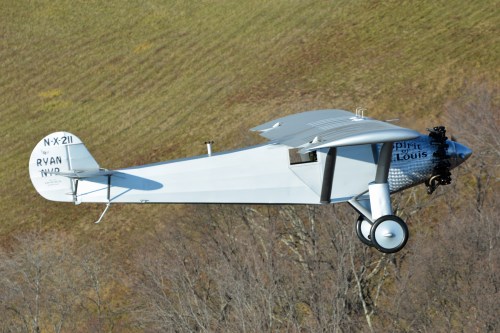 A vintage airplane in flight, showcasing a sleek design and a silver body against a green landscape.