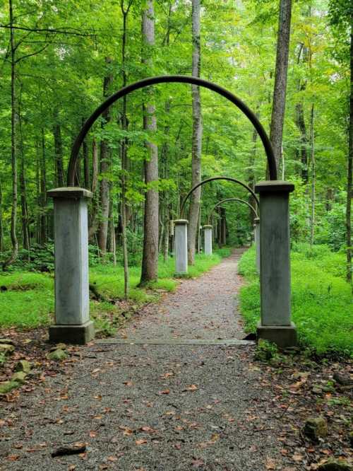 A serene forest path lined with archways, surrounded by lush green trees and a gravel walkway.