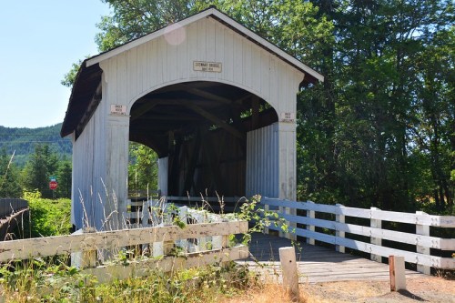 A white wooden covered bridge with a sign, surrounded by greenery and a wooden fence.
