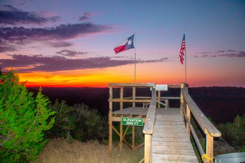 A wooden observation deck with Texas and American flags at sunset, overlooking a scenic landscape. Elevation sign: 2002 ft.