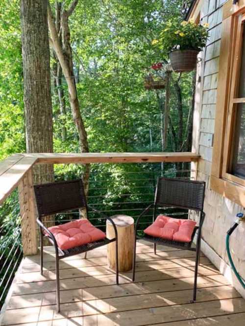 Two chairs with cushions and a small wooden table on a wooden deck surrounded by lush green trees.