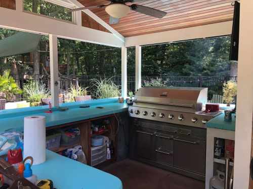 A modern outdoor kitchen with a stainless steel grill, turquoise countertops, and a ceiling fan in a wooded setting.