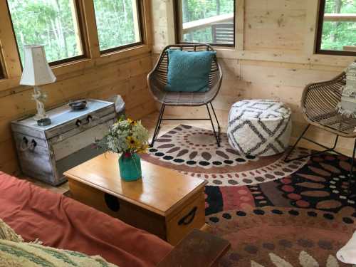 Cozy interior with a wooden floor, a sofa, decorative pouf, and a table with flowers in a vase, surrounded by windows.