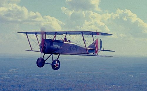 A vintage biplane flying high above a landscape with clouds in the background.