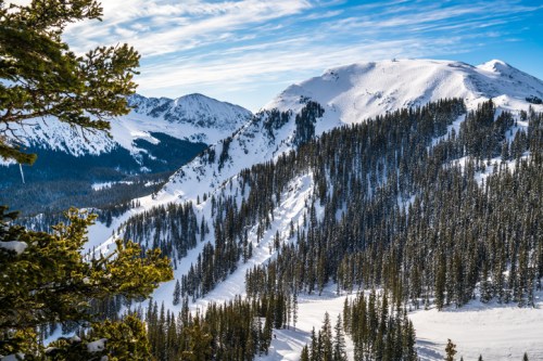 Snow-covered mountains under a blue sky, surrounded by evergreen trees, creating a serene winter landscape.