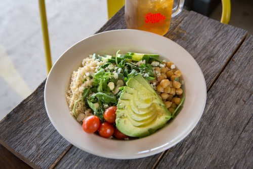 A bowl of quinoa, chickpeas, cherry tomatoes, avocado, and greens on a wooden table, with a drink in the background.
