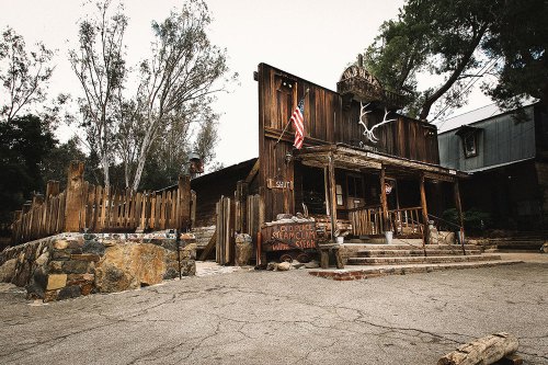 A rustic wooden building with an American flag, surrounded by trees and a stone pathway, evoking a western theme.