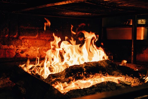 A close-up of a roaring fire with bright flames and glowing embers in a dark, rustic setting.