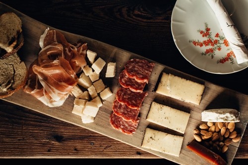 A wooden platter with assorted cheeses, cured meats, bread, almonds, and a decorative plate in the background.