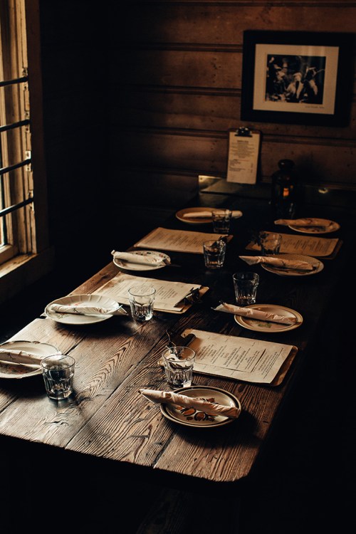 A rustic dining table set with plates, glasses, and menus, illuminated by natural light from a nearby window.