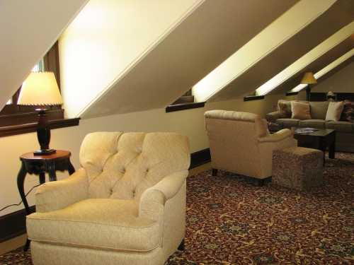 Cozy attic lounge with sloped ceilings, featuring beige armchairs, a side table, and a sofa on patterned carpet.