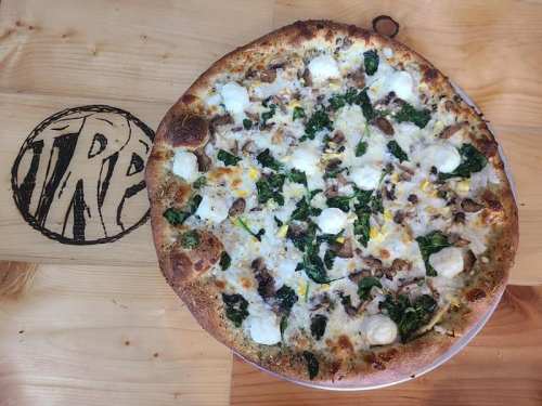 A freshly baked pizza topped with spinach, cheese, and chicken, served on a wooden table with a logo.