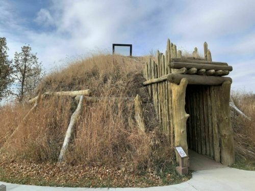 A grassy, earthen mound with a wooden entrance, surrounded by tall grass and trees under a cloudy sky.