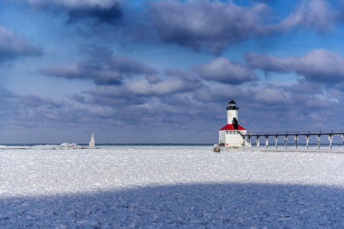 A lighthouse stands on a frozen shoreline under a cloudy sky, with ice covering the ground and water.