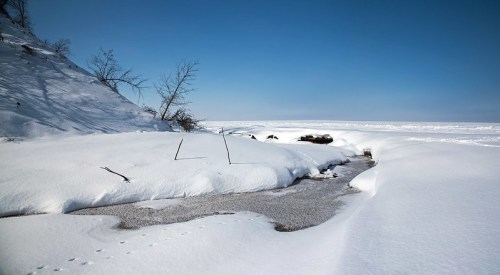 A snowy landscape with a frozen lake, bare trees, and a clear blue sky.