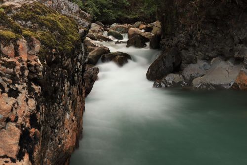 A serene river flows between rocky banks, with smooth water creating a misty effect in a lush, green landscape.