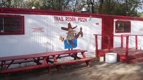 A colorful mural of a cowboy with a horse on the exterior of Trail Rider Pizza, featuring red picnic tables outside.