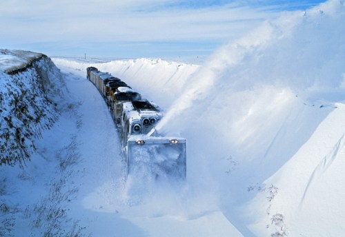 A snowplow train clears a path through deep snow, surrounded by high snowbanks under a clear blue sky.