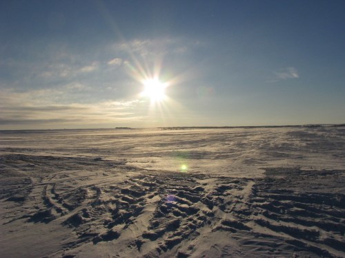A vast, snowy landscape under a bright sun, with clear blue skies and distant horizon.