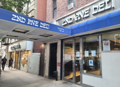 Exterior of 2nd Ave Deli with blue awning, featuring storefront and sidewalk with pedestrians.