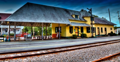 A yellow train station with a sloped roof, surrounded by tracks and greenery under a blue sky.