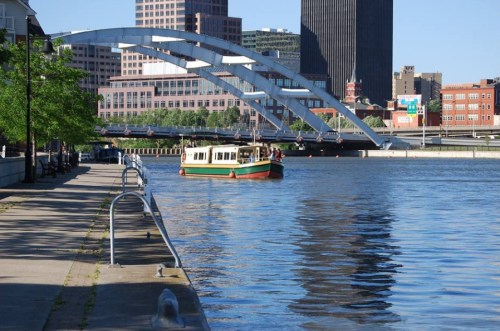 A boat glides on a river with a city skyline and a large arch bridge in the background on a sunny day.
