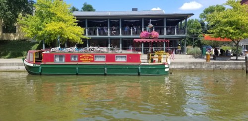 A colorful houseboat docked by a waterfront restaurant with green trees and a clear blue sky in the background.