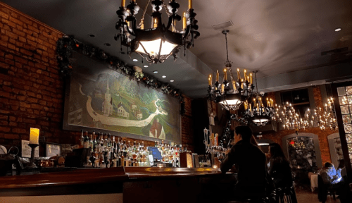 Cozy bar interior with brick walls, chandeliers, and a mural, featuring a person sitting at the counter.