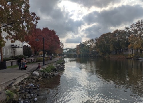 A serene river scene with autumn trees, a walkway, and people sitting by the water under a cloudy sky.
