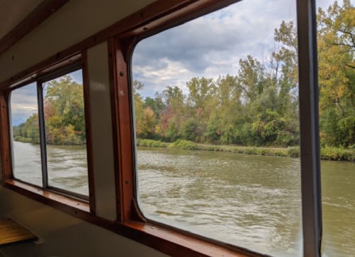 View from a boat window showing a calm river and trees with autumn foliage along the banks.