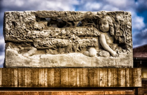 A carved stone relief depicting a child among floral designs, set against a cloudy sky.