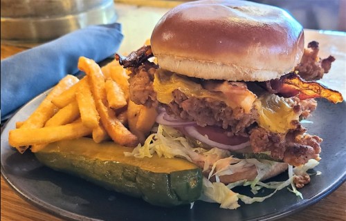 A crispy chicken sandwich with cheese, bacon, lettuce, and onions, served with fries and a pickle on the side.