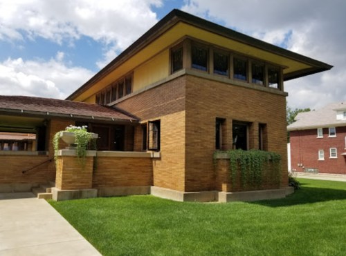 A two-story brick building with large windows, surrounded by green grass and a cloudy sky.