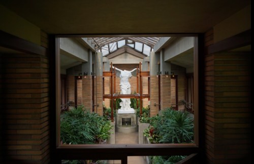 A view through a window framing a statue surrounded by lush greenery in a modern architectural space.