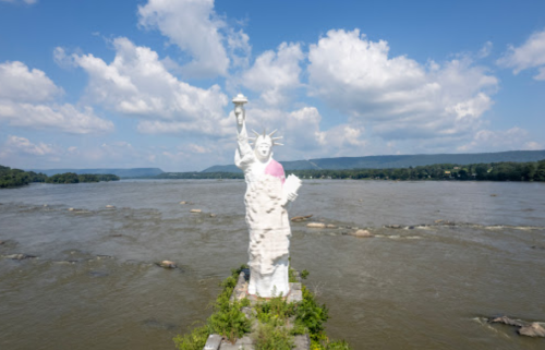 A large statue resembling the Statue of Liberty stands on a rocky riverbank under a blue sky with fluffy clouds.