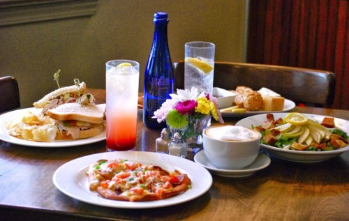 A table set with a sandwich, pizza, salad, drink, and dessert, surrounded by flowers and water bottles.