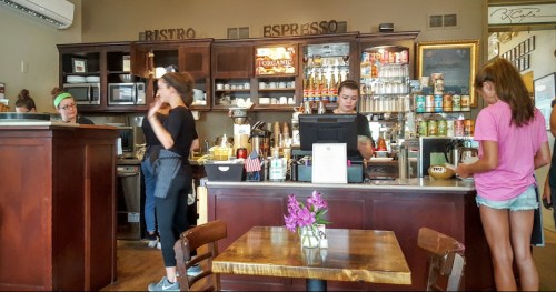 A cozy café interior with a counter, baristas serving customers, and a table with flowers in the foreground.