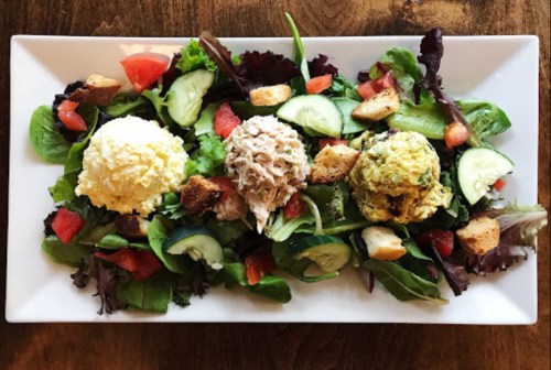 A platter of mixed greens topped with three scoops of salad: chicken, egg, and tuna, garnished with tomatoes and cucumbers.