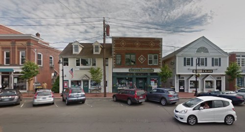 A street view of a small town with shops, parked cars, and a cloudy sky.