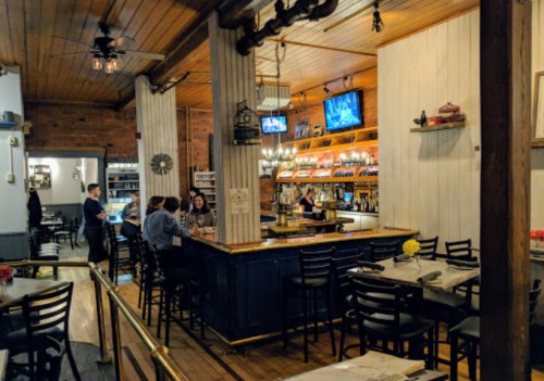 Cozy restaurant interior with a bar, wooden decor, and patrons enjoying their meals at tables and the bar.