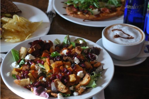 A colorful salad with greens, roasted vegetables, croutons, and goat cheese, alongside a cappuccino and chips.