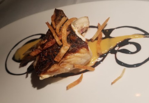 A piece of grilled fish topped with crispy strips and drizzled with a yellow sauce on a decorative plate.