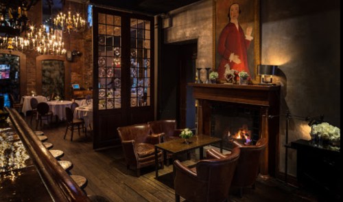 Cozy restaurant interior with a fireplace, elegant seating, and a large painting on the wall, illuminated by chandeliers.
