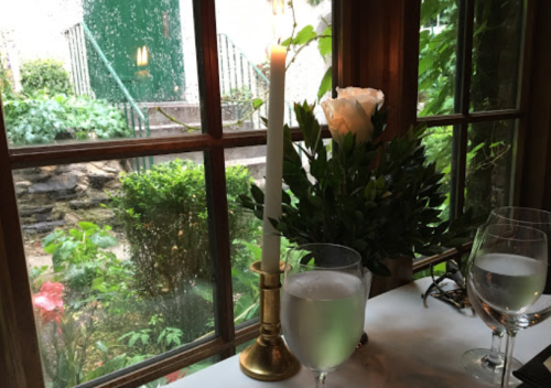 A cozy table setting with a candle, a glass of water, and a white rose, viewed through a rain-speckled window.