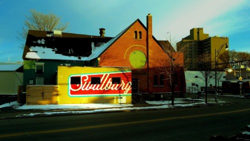 A colorful mural reading "Sudbury" on a building, with snow on the ground and city buildings in the background.