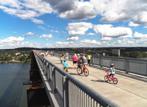 A busy bridge with people walking and biking, surrounded by blue skies and clouds, overlooking a river and landscape.
