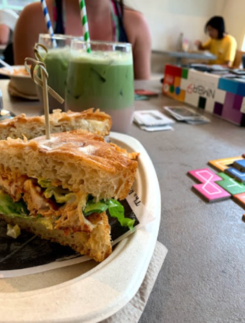A close-up of a sandwich with greens, alongside two drinks and a board game in a casual dining setting.